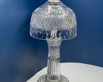 May include: A clear glass table lamp with a diamond-cut pattern on the shade and base. The lamp has a white cord and plug. The lamp is on a white surface with a blue background.