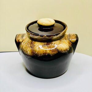 May include: A brown and cream glazed ceramic cookie jar with a lid. The jar has two handles on the sides and a round lid with a knob.