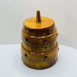 May include: A ceramic cookie jar shaped like a wooden barrel with a lid. The jar is brown and yellow with a wood grain pattern and has a pointed lid.