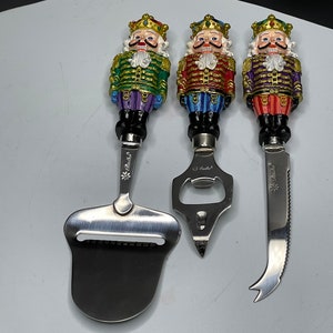 May include: Set of three nutcracker themed kitchen tools. A cheese grater, bottle opener, and butter knife. Each tool has a different colored nutcracker handle with a gold crown.