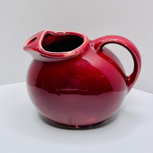 May include: A deep red ceramic pitcher with a rounded body and a single handle. The pitcher has a smooth, glossy finish.