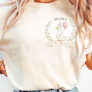 May include: Cream-coloured t-shirt featuring a cartoon goose wearing a party hat and holding a pink balloon. The text "MAMA of the silly goose" is above and below the goose illustration. The shirt has rolled-up sleeves and is a relaxed fit.