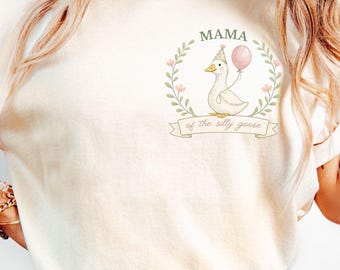 May include: Cream-coloured t-shirt featuring a cartoon goose wearing a party hat and holding a pink balloon. The text "MAMA of the silly goose" is above and below the goose illustration. The shirt has rolled-up sleeves and is a relaxed fit.