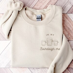 May include: A beige sweatshirt with the text "In my sourdough era" and a graphic of four jars of sourdough starter.