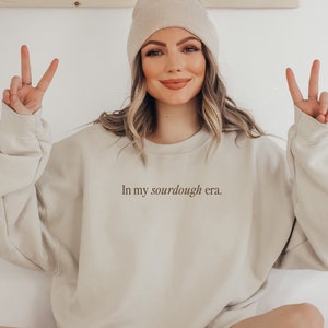 May include: A woman wearing a beige sweatshirt with the text "In my sourdough era." printed on the front. She is wearing a beige beanie and has her hands up in a peace sign.