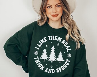 Può includere: Una felpa verde scuro con una stampa grafica bianca che dice "Mi piacciono veri, spessi e profumati di pino" con un'immagine stilizzata di tre alberi di Natale. La felpa è indossata da una persona con un cappello bianco.