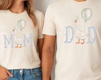 May include: Matching beige t-shirts featuring a goose illustration with a party hat and balloon. One shirt says "MOM" in light blue, the other "DAD". Both people are wearing blue jeans. The shirts are a fun, coordinated outfit.