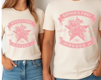 May include: Two cream-coloured t-shirts with a pink graphic. The graphic features the text "SOPHIA'S FIRST" in a banner, a star, a horse and rider, and the text "AND 20 RODEO 24". The shirts are paired with blue jeans.