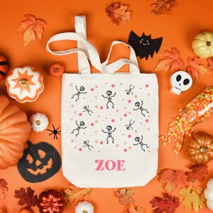 Puede incluir: Una bolsa de lona blanca con el nombre "ZOE" impreso en rosa en la parte delantera. La bolsa está decorada con un patrón repetido de esqueletos negros y flores rosas. La bolsa está rodeada de varias decoraciones de Halloween, incluyendo calabazas, maíz dulce y hojas de otoño.