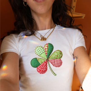 Może przedstawiać: Biała koszulka z dużym, kolorowym wzorem koniczyny. Koniczyna wykonana jest z zielonej i czerwonej tkaniny z różnymi wzorami, w tym grochami, kratką i nadrukami kwiatowymi. Codzienny, swobodny element garderoby.