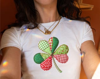 May include: White t-shirt with a large shamrock design. The shamrock is made of green and red patterned fabric, including polka dots, gingham, and floral prints. A casual, everyday wear item.