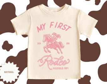 May include: A cream-coloured t-shirt with the text "MY FIRST Rodeo" in pink, featuring a pink graphic of a person riding a horse. The shirt also includes the text "SADDLE UP!" and the numbers "20" and "26".