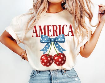 May include: A white t-shirt with the word "AMERICA" printed in red letters. A blue and white bow with stars is above two red cherries with white stars.