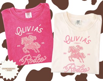 May include: Two t-shirts, one in pink and one in ivory, with a rodeo design. Each shirt displays the text "Olivia's First Rodeo" and a graphic of a person riding a horse. The shirts are labelled "Crunchberry + Ivory".