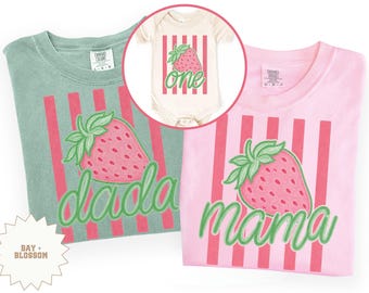 May include: A light green t-shirt with a strawberry graphic and the word "dada" in green script. A pink t-shirt with a strawberry graphic and the word "mama" in green script. A cream-coloured babygrow with a strawberry and the word "one".