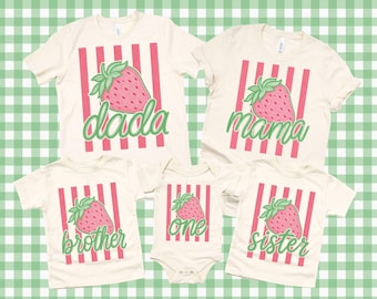 May include: A collection of five cream-coloured shirts and one bodysuit featuring a graphic of a strawberry over vertical pink stripes. The garments include text labels in green cursive script reading dada, mama, brother, one, and sister.