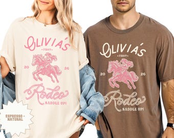 May include: Two t-shirts, one cream and one brown, displaying a pink rodeo design. The design includes the text "Olivia's First" above a pink illustration of a cowgirl riding a horse, with "20 26" on either side. Below, the word "Rodeo" and "Saddle Up!" are written in a decorative font.