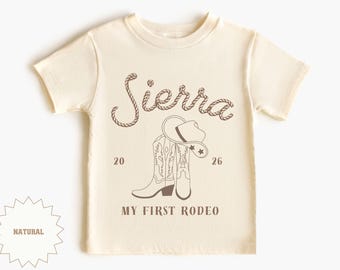 May include: Cream-coloured t-shirt with a brown graphic. The design includes the name "Sierra" in a rope-style font, cowboy boots, a hat, and the text "MY FIRST RODEO". The numbers "20" and "26" are also present.