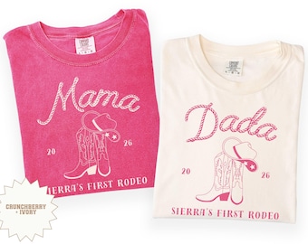 May include: Two t-shirts, one cerise and one cream, with rodeo-themed graphics. The cerise shirt says "Mama" and the cream shirt says "Dada" in a rope-style font. Each shirt features a cowboy hat and boots, with "Sierra's First Rodeo" printed below.