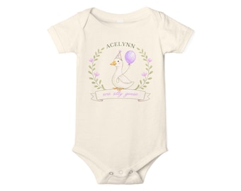 May include: A cream-coloured baby bodysuit featuring a cartoon goose wearing a party hat and holding a purple balloon. The design includes the name "ACELYNN" and the phrase "one silly goose" within a floral wreath.
