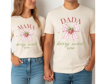 May include: Two cream-coloured t-shirts with pink and white gingham bows and strawberries. The text on the shirts reads "Mama of the berry sweet one" and "Dada of the berry sweet one".