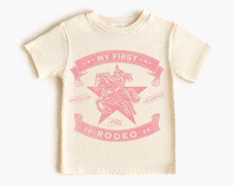 May include: A cream-coloured t-shirt with a pink graphic. The graphic shows a cowgirl riding a horse, a star, and the text "MY FIRST" above and "RODEO 26" below. The shirt is a short-sleeved crew neck.