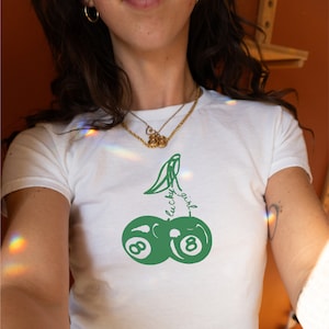 Op de afbeelding: Wit T-shirt met een groene afbeelding van een kers met de woorden "lucky girl" op de steel. De kers is gemaakt van twee groene biljartballen met het nummer 8 op elk.