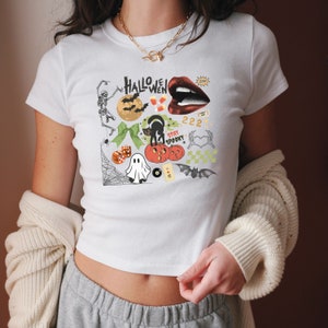 May include: White cropped t-shirt with a Halloween themed graphic print. The print features a variety of spooky and festive elements, including a ghost, a black cat, a spider web, dice, pumpkins, candy corn, and the text "HALLOWEEN".