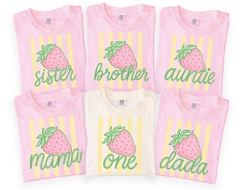 May include: Six short-sleeved t-shirts, five pink and one cream. Each has a strawberry graphic and text: "sister", "brother", "auntie", "mama", "one", and "dada". Yellow vertical stripes are behind the strawberry design on each shirt.