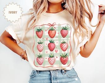 May include: A white t-shirt featuring a graphic of strawberries on a green and white gingham pattern. The strawberries are red with green leaves and are arranged in a square shape. The shirt has a bow at the top of the graphic.