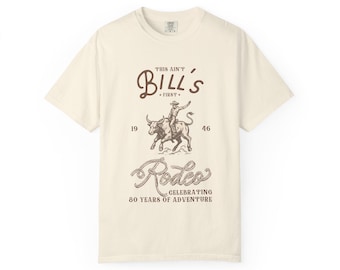 May include: Off-white t-shirt with a rodeo-themed design. The graphic depicts a cowboy on a bull, with the text "This Ain't Bill's First," and "Rodeo Celebrating 80 Years of Adventure." The shirt has short sleeves and a round neck.