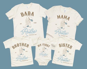 May include: Off-white family matching t-shirts and a onesie with a rodeo theme. The shirts say "Dada", "Mama", "Brother", and "Sister", and the onesie says "My First Rodeo". Each has a cowboy boot and hat graphic.