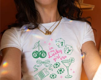 May include: White t-shirt with a graphic design featuring cherries, dice, casino chips, a cocktail, and the text "in my Lucky girl era" in pink and green. The design includes a bow and a ticket.