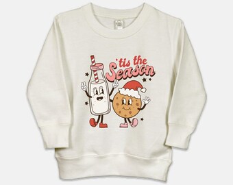 May include: A white long-sleeved sweatshirt featuring a cartoon illustration of a milk bottle and a cookie wearing a Santa hat. The text 'tis the season is printed in red and pink on the sweatshirt.
