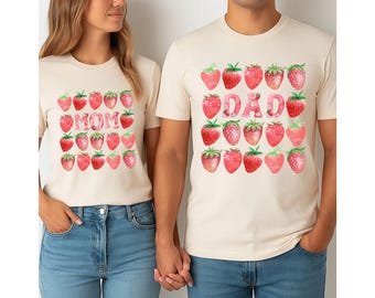 May include: Matching cream-coloured t-shirts with strawberry designs. One shirt says "MOM" in pink letters, the other says "DAD". Both shirts have rows of red strawberries. The people are wearing blue jeans.