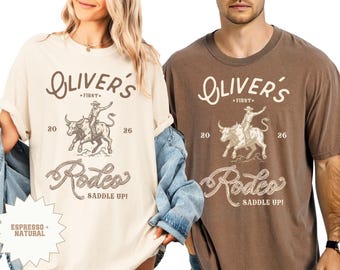 May include: Two t-shirts, one cream and one brown, with a rodeo design. The design includes the text "Oliver's Rodeo" with a bull rider graphic and the words "Saddle Up!"