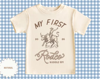 May include: A cream-coloured t-shirt featuring a brown graphic of a cowboy riding a bull. The text "MY FIRST Rodeo" is arched above the image, with "20 26" on either side. The words "SADDLE UP!" are below the image.