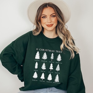 Pode incluir: Uma camisola verde escura com um gráfico branco de diferentes tipos de árvores de Natal e seus nomes. O texto diz "O Christmas Tree" e inclui os nomes das árvores: "Grand Fir", "Douglas Fir", "Lodge Pole", "Virginia Pine", "Hemlock", "Noble Fir", "White Fir", "Silver Tip", "Fraser Fir", "White Pine", "Balsam Fir", "Norway Spruce".