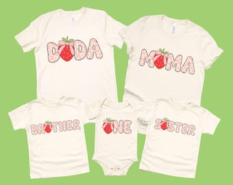 May include: Cream-coloured t-shirts and a onesie with strawberry-themed designs. The shirts display the words "DODA", "MOMA", "BROTHER", "ONE", and "SISTER" in a floral font, each paired with a strawberry graphic. The background is a light green.