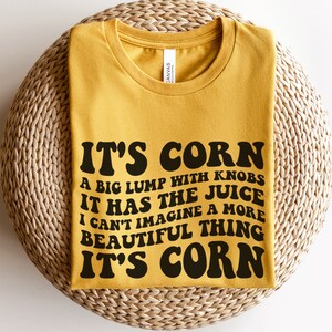 Può includere: Una maglietta gialla con testo nero che recita "It's Corn. A big lump with knobs. It has the juice. I can't imagine a more beautiful thing. It's Corn."