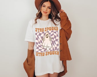May include: White t-shirt with a graphic of a ghost with the text "Stay Spooky" in a retro font. The ghost is on a purple and white checkered background.