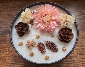 May include: A white candle with a black lid, decorated with dried flowers, pine cones, and gold glitter.