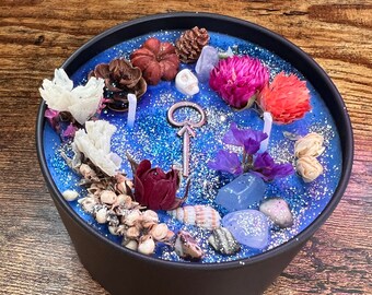May include: A black candle holder filled with blue wax and decorative elements. The candle is adorned with dried flowers, crystals, seashells, and a key-shaped charm. The arrangement includes a variety of colors and textures, creating a unique aesthetic.