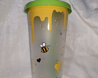 May include: A clear reusable cup with a yellow and green lid and a green straw. The cup has a yellow drip design and is decorated with a bee, hearts, and confetti.