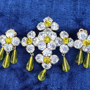 May include: A vintage jewelry set featuring a central brooch and matching earrings. The pieces are adorned with clear and yellow rhinestones arranged in a floral pattern, with dangling teardrop-shaped green accents. The set is set against a blue background.