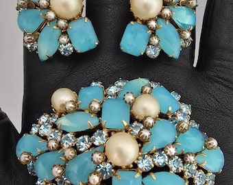May include: A vintage brooch and earring set. The jewellery features light blue, pearlescent, and clear gemstones set in a gold-tone metal. The earrings are clip-on style. The brooch is a large, eye-catching piece.