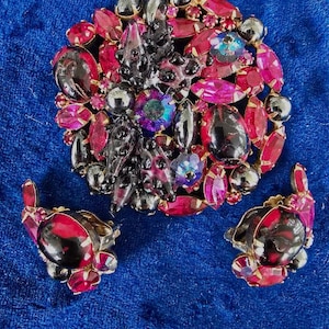 May include: A vintage brooch and earrings set featuring a cluster of dark red, pink, and iridescent rhinestones with a black metal base.