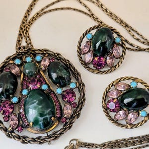 May include: A vintage jewellery set comprising a pendant necklace and matching clip-on earrings. The set features a large pendant with a dark green oval stone, surrounded by pink and turquoise stones, and a gold-tone metal frame. The earrings have a similar design.