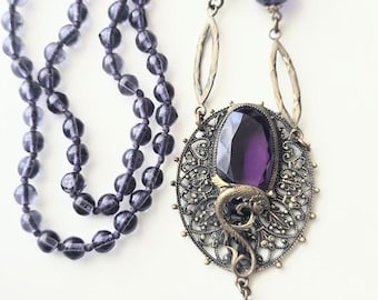May include: A vintage necklace with a large, oval purple gemstone pendant, ornate metal filigree, and a small snake detail. The necklace has strands of small, round, purple beads. The pendant is suspended from a chain.