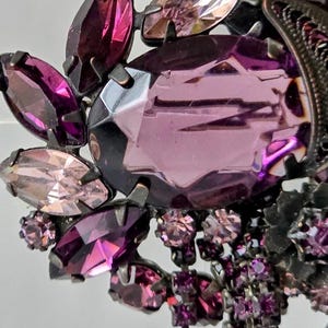 May include: A vintage brooch with a large, faceted, oval purple gemstone, surrounded by smaller, marquise-cut and round purple and pink crystals. The brooch has a dark metal setting and dangling crystal accents, creating a glamorous, ornate design.
